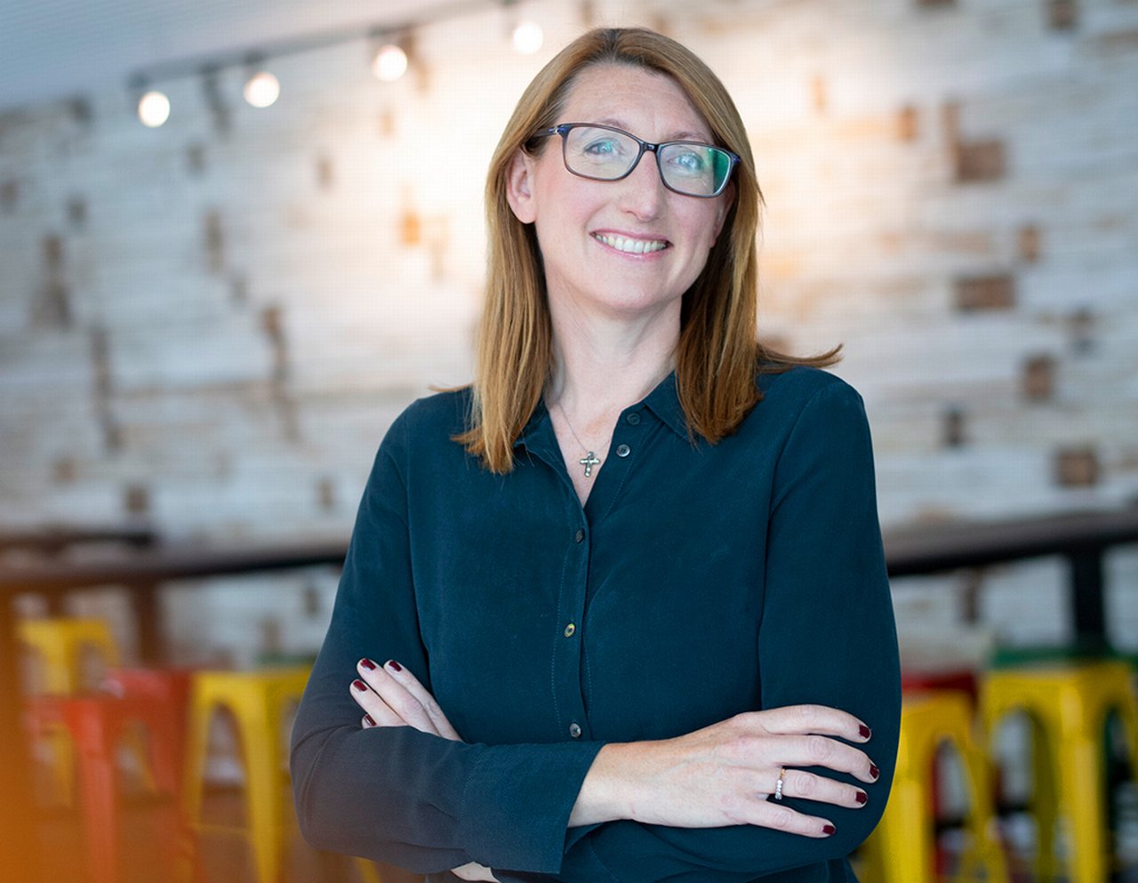 Deirdre is standing in the canteen with her arms crossed smiling