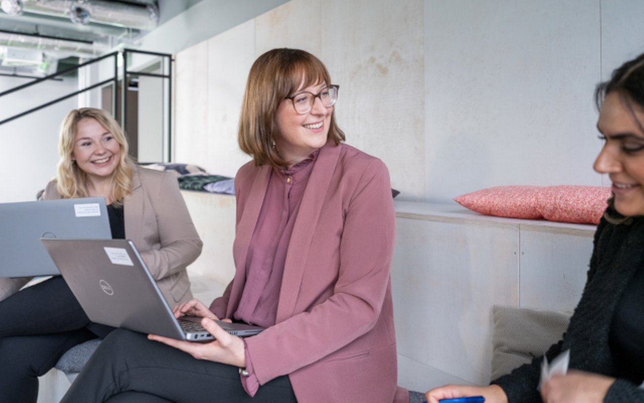 Drei Personen in einem modernen Büro, zwei arbeiten am Laptop, eine Person macht sich Notizen; im Hintergrund industrielle Deckenelemente.